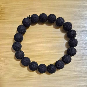 May include: A circular bracelet composed of twenty-one individual black lava stone beads. Each bead features a porous, textured surface and a matte finish. The beads are strung together in a continuous loop, with a small black knot visible at the top.