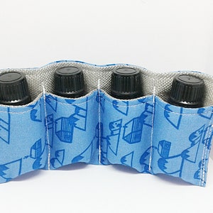 May include: A light blue essential oil bottle holder with four compartments. The holder is made of fabric with a blue and white geometric pattern. Each compartment holds a black bottle with a black cap. The interior lining is gray.