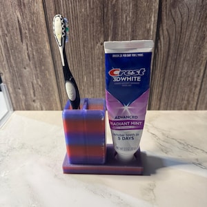 May include: A 3D-printed toothbrush holder in shades of purple and red, holding a black toothbrush and a tube of Crest 3D White toothpaste. The toothpaste tube is white with blue and purple accents, and the text "ADVANCED RADIANT MINT" is visible.