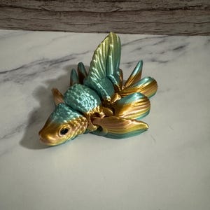 May include: A decorative, 3D-printed fish figurine with a teal and gold color scheme. The fish has detailed scales and fins, and is posed as if swimming. The figurine is approximately 4 inches long.