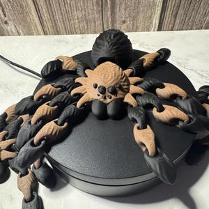 May include: A 3D-printed spider sculpture with a black body and legs, and a tan-colored head and abdomen. The spider is posed on a black circular base, with its legs arranged around the base. The sculpture is likely a decorative item or a unique piece of art.