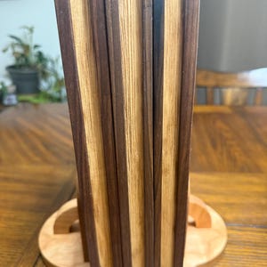 May include: A wooden book stand with a light-colored base and a vertical support made of alternating dark and light wood strips. The stand is designed to hold books or display items, showcasing a natural wood grain pattern.