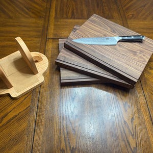 May include: A wooden knife block and three wooden cutting boards are displayed on a wooden table. A stainless steel knife with a black handle rests on top of the cutting boards. The knife block is light-colored wood.