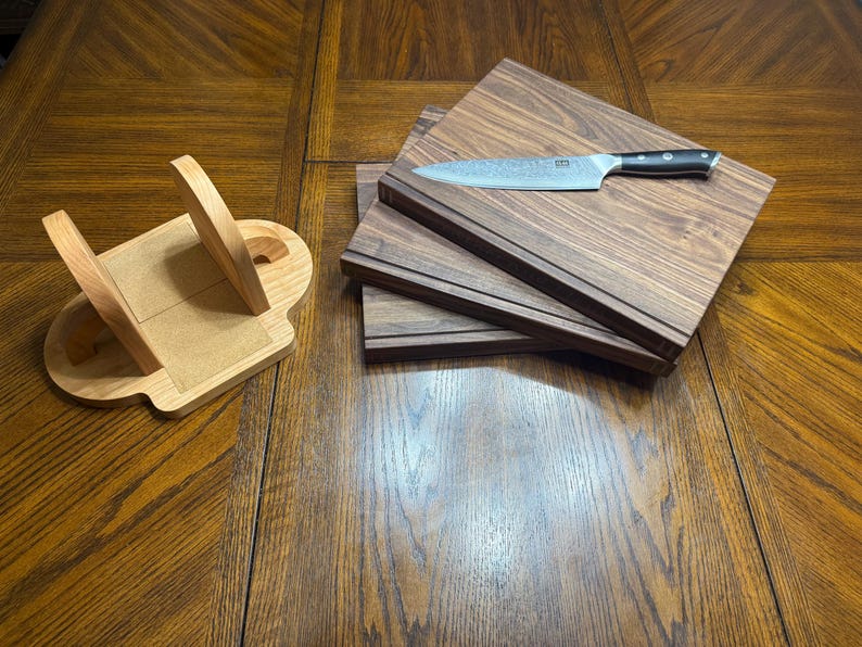 May include: A wooden knife block and three wooden cutting boards are displayed on a wooden table. A stainless steel knife with a black handle rests on top of the cutting boards. The knife block is light-colored wood.