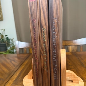 May include: Three hardcover books with dark wood covers are displayed in a wooden bookend. The book spines feature titles in a handwritten style with red, green, and blue accents. The bookend is made of light-colored wood.