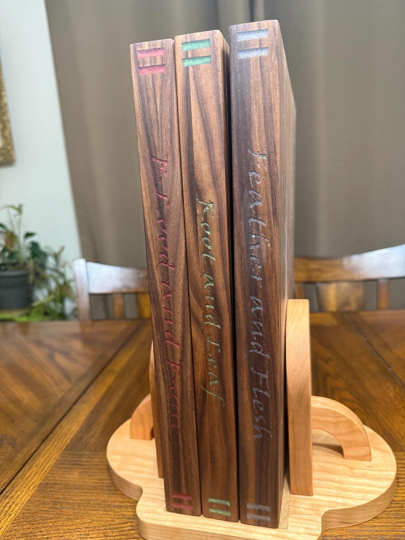May include: Three hardcover books with dark wood covers are displayed in a wooden bookend. The book spines feature titles in a handwritten style with red, green, and blue accents. The bookend is made of light-colored wood.
