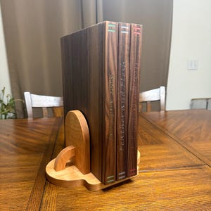 May include: A wooden bookend set with a light brown base and a dark brown book holder. Three books with the titles "Book 1", "Feather and Flesh", and "Local and Lost" are held in place. The bookends are on a wooden table.