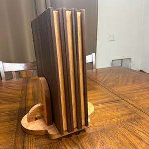 May include: A wooden coaster set with a holder. The coasters are made of alternating dark and light wood, creating a striped effect. The holder is a light-colored wood with a curved design, providing a stylish way to store the coasters. The set is displayed on a wooden table.