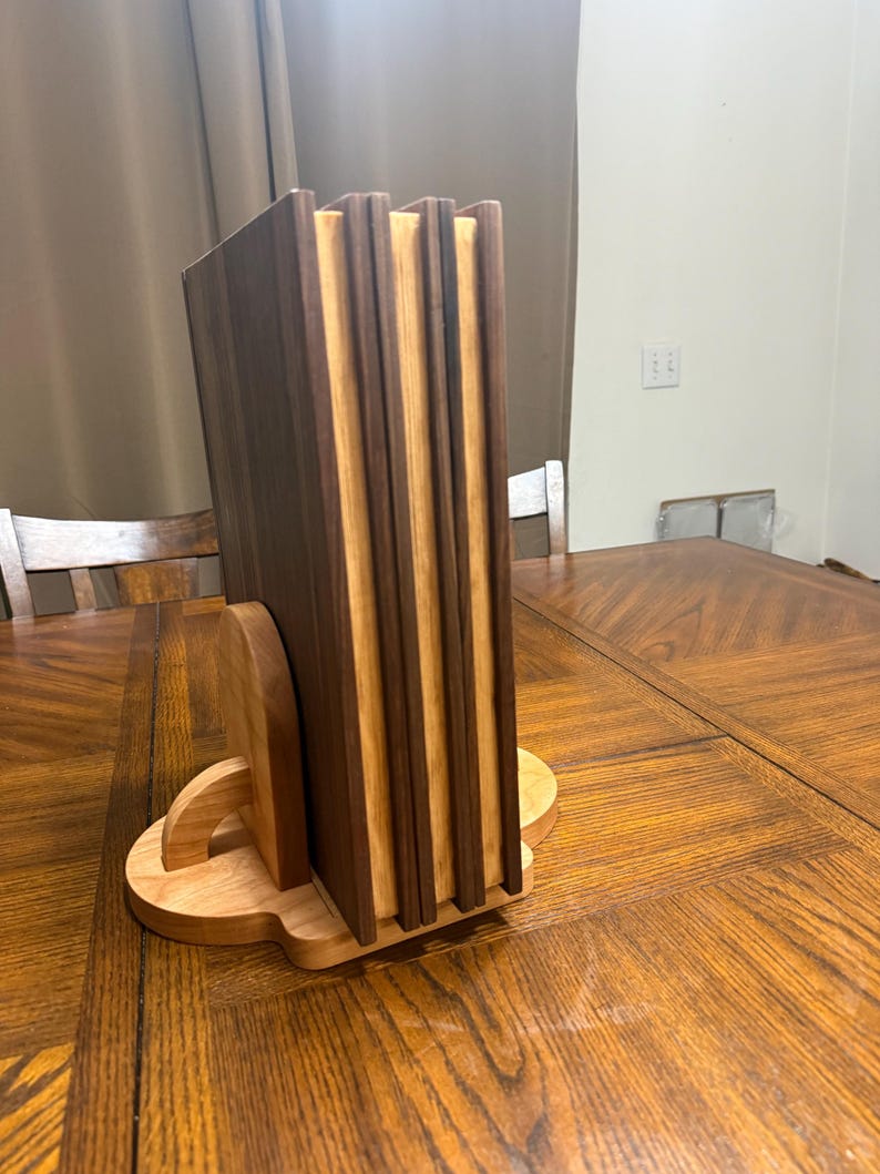 May include: A wooden coaster set with a holder. The coasters are made of alternating dark and light wood, creating a striped effect. The holder is a light-colored wood with a curved design, providing a stylish way to store the coasters. The set is displayed on a wooden table.