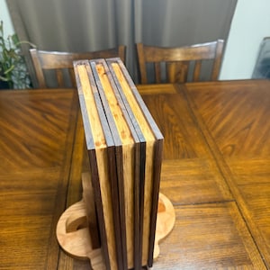 May include: A wooden book stand with a stack of books. The books have alternating dark and light wood stripes. The stand is made of light-colored wood and is on a wooden table.