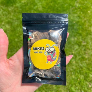 May include: A black, resealable bag filled with dark brown, dried food items. The bag has a yellow circular label with the text "MIKES BUG BOX" and a cartoon bug illustration. The bag is held against a green, grassy background.