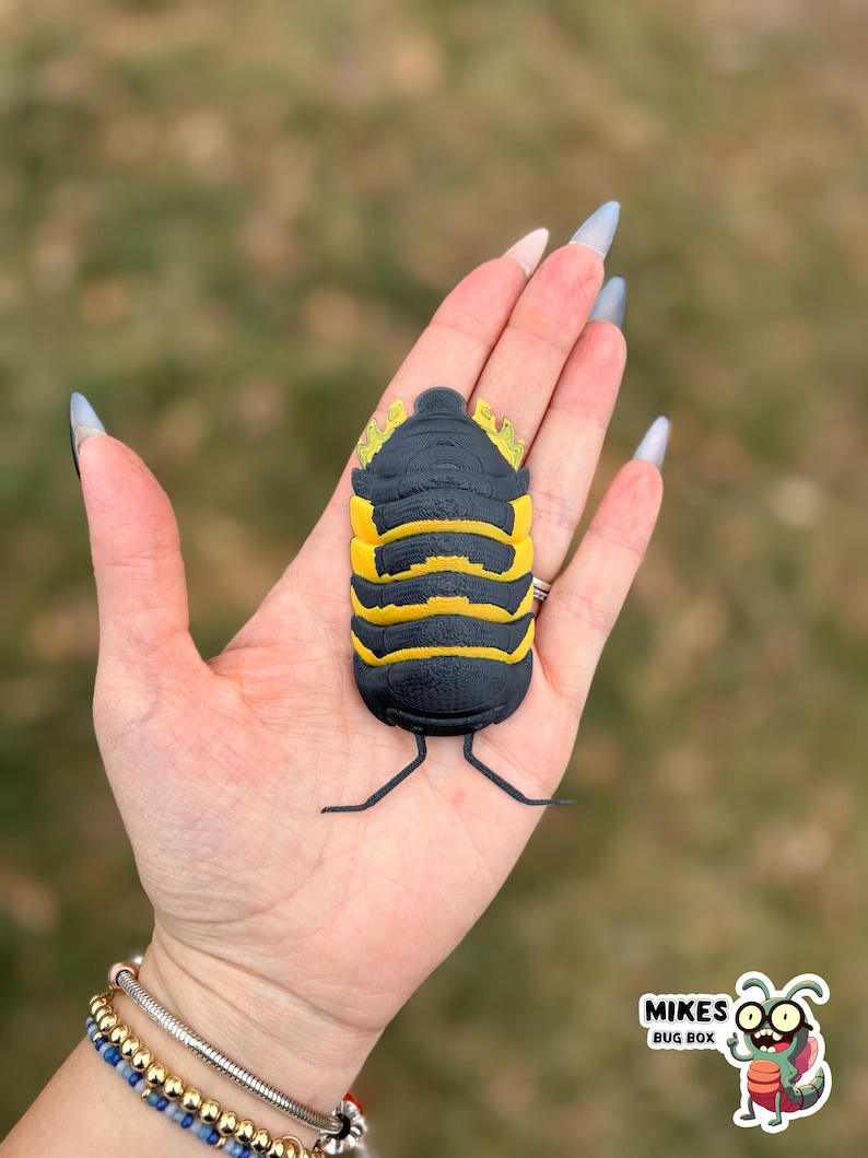 May include: A black and yellow isopod bug figurine with a segmented body and small legs. The figurine is held in a hand against a blurred green background. The figurine has a detailed design and is a unique decorative item.