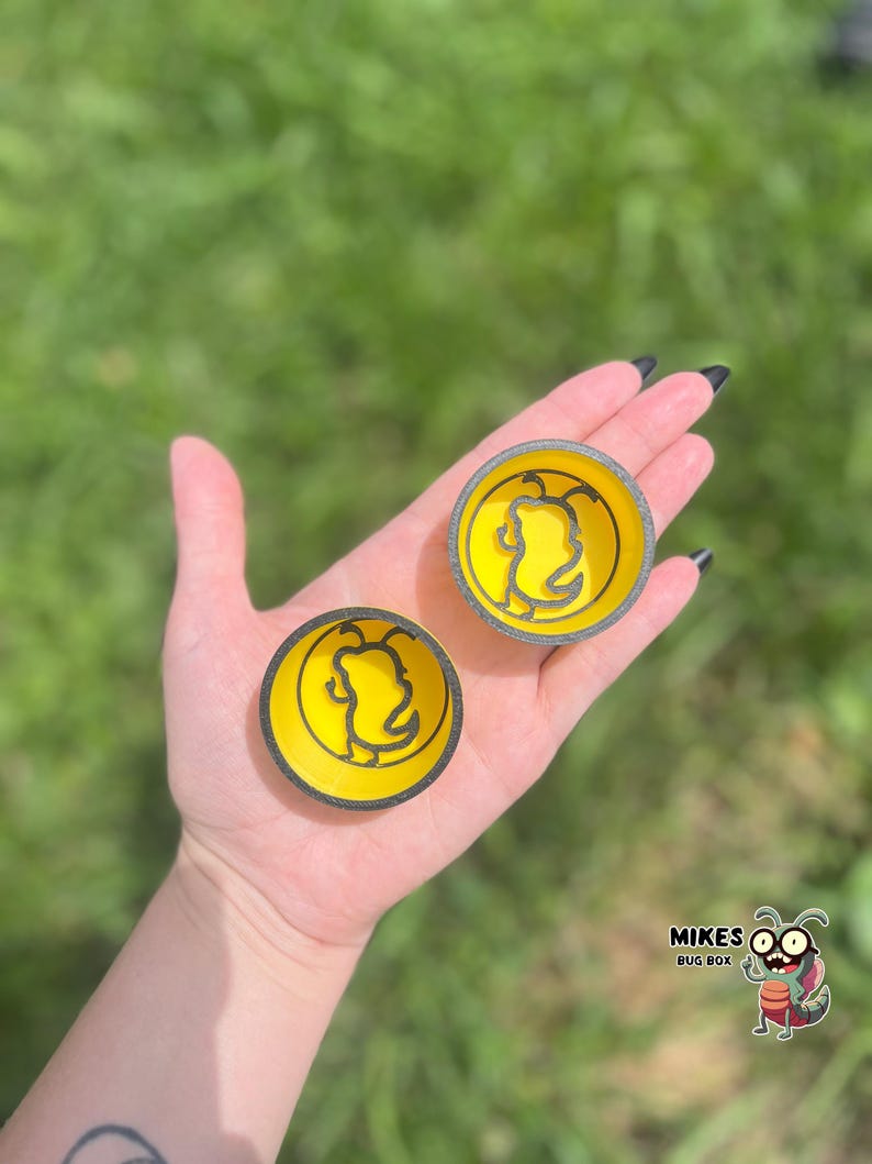 May include: Two yellow and black circular objects with a bee design are held in a hand. The objects have a yellow center with a black outline of a bee. The outer rim is black. The objects are likely coasters or small decorative items. The logo "MIKES BUG BOX" is in the bottom right corner.