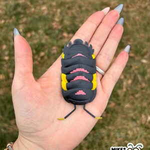 May include: A 3D-printed bug toy with a segmented black body, yellow accents, and pink details. The toy is held in a hand with long, gray fingernails. The logo "MIKES BUG BOX" is visible in the lower right corner.