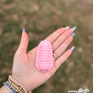May include: A pink, segmented toy bug with small black eyes is held in a hand. The toy is a light pink color and has a ridged texture. The hand is adorned with bracelets and rings. The background is blurred, with a logo in the bottom right corner.