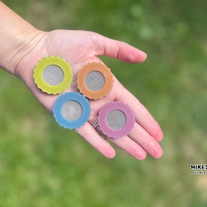 Puede incluir: Cuatro pantallas de repuesto circulares y coloridas para cajas de insectos en amarillo verdoso, naranja, azul y rosa. Cada pantalla tiene un centro de malla y un borde festoneado. Las pantallas se sostienen en una mano sobre un fondo verde borroso.