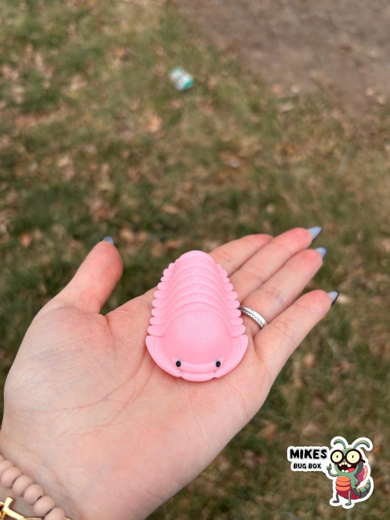 May include: A pink trilobite toy with small black eyes is held in a hand. The toy is a light pink color and has a segmented body. The background is blurred, showing grass and a small amount of trash. The image includes a logo for "Mikes Bug Box."