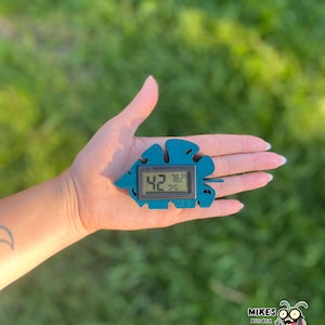 May include: A teal leaf-shaped digital hygrometer displaying 42% and 78.2°F. The device has a small LCD screen and the text "M.B.C." The background is a blurred green lawn.