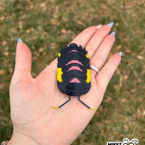 May include: A black, yellow, and pink bug-shaped figurine with thin yellow legs. The figurine is held in an open hand against a blurred green background. The figurine has a segmented body and is likely a decorative item.
