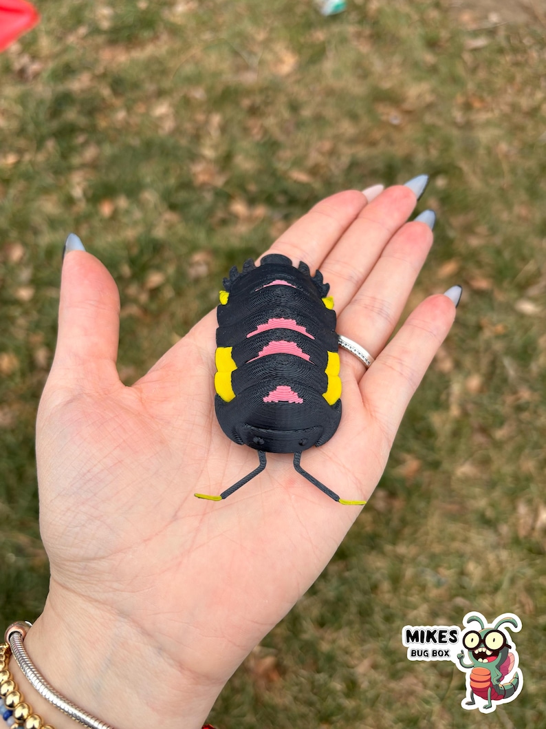 May include: A black, yellow, and pink bug-shaped figurine with thin yellow legs. The figurine is held in an open hand against a blurred green background. The figurine has a segmented body and is likely a decorative item.