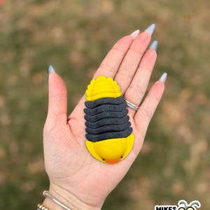 May include: A yellow and black bug-shaped figurine with a segmented body and a smiling face. The figurine is held in a hand, and the background is blurred. The figurine has the text "MIKES BUG BOX" on the bottom right.