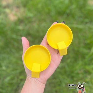 May include: Two bright yellow, circular containers with rectangular tabs are held in a hand. The containers are open, revealing their interior. The background is blurred green grass. The image includes the text "MIKES BUG BOX" with a cartoon bug.