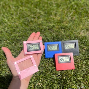 May include: A hand holding several small, colorful digital hygrometers. The hygrometers are pink, blue, red, and black, displaying temperature and humidity readings. One pink frame is also visible. The grass is green.