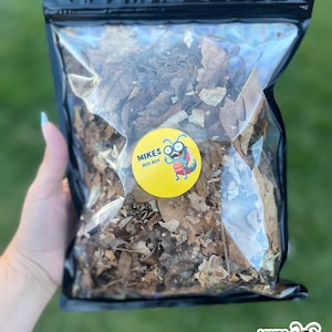 May include: A clear plastic bag filled with brown, dry, shredded leaves. The bag has a black zipper closure and a yellow logo that says "Mikes Bug Box" with a cartoon bug wearing glasses.