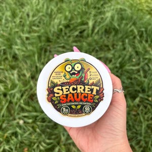 Puede incluir: Un recipiente blanco con las palabras "SECRET SAUCE" en letras grandes. El recipiente tiene un dibujo animado de un insecto verde con ojos grandes. También es visible el texto "BOOST YOUR SUBSTRATE. FEED YOUR GROW". El recipiente contiene 142 gramos.