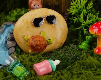 May include: A miniature scene with a brown rock wearing sunglasses, a small turtle figurine, a green snake figurine, a red and white mushroom figurine, a clear glass with a green liquid, and a pink baby bottle.