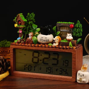 May include: A wooden digital clock with a brown wood frame and a white digital display showing the time 8:23. The clock is decorated with miniature figurines of animals and plants, including a birdhouse, a squirrel, an owl, and a small tree.