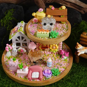 Könnte beinhalten: Ein zweistöckiges Holztablett mit einem rosa-weißen Miniaturhaus, einer Hasenfigur, einer kleinen Bank und einer Vielzahl anderer Miniaturdekorationen. Das Tablett ist mit rosa-weißen Streuseln verziert und trägt ein Schild mit der Aufschrift "Park".