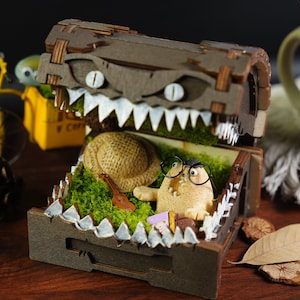 May include: A wooden treasure chest with a toothy monster face on the lid is open, revealing a small, brown, furry creature wearing glasses and holding a guitar. The chest is filled with green moss and a brown hat.
