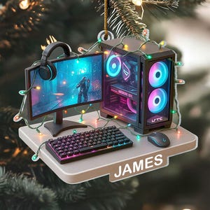 May include: A Christmas ornament featuring a miniature gaming setup. It includes a monitor, keyboard, mouse, and a PC tower with colorful lights. The name "JAMES" is printed on the base. The ornament is decorated with string lights and a headset.