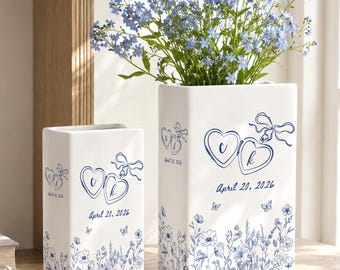 May include: Two white rectangular ceramic vases featuring a blue line art design of two heart-shaped lockets with the letters C and K, a ribbon, and the date April 20, 2026. The base of each vase is decorated with a blue floral and butterfly pattern.