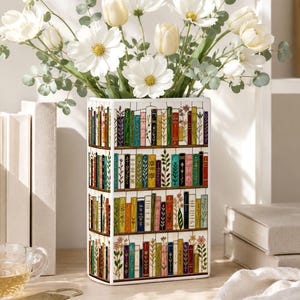 May include: A rectangular ceramic vase featuring a multi-tiered bookshelf illustration with colorful book spines and floral accents. The vase is filled with a bouquet of white cosmos flowers and green eucalyptus stems, standing upright on a flat surface.