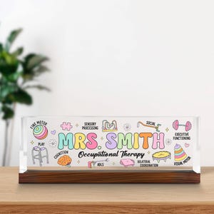 May include: A rectangular clear acrylic desk sign with a dark wood base featuring the text MRS. SMITH Occupational Therapy in colorful bubble letters, surrounded by illustrations of therapy tools like a walker, brain, and puzzle piece.