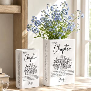 May include: Two white rectangular ceramic vases featuring black text that reads New Chapter and a floral illustration. The larger vase contains a bouquet of small blue flowers. The text includes a quote and the name Jenifer printed on the front.