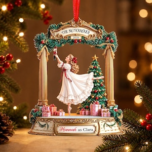May include: A decorative Christmas ornament featuring a ballerina in a white dress holding a nutcracker under an archway. The arch is inscribed with THE NUTCRACKER 2026, and the base displays the text Hannah as Clara, surrounded by gift boxes.