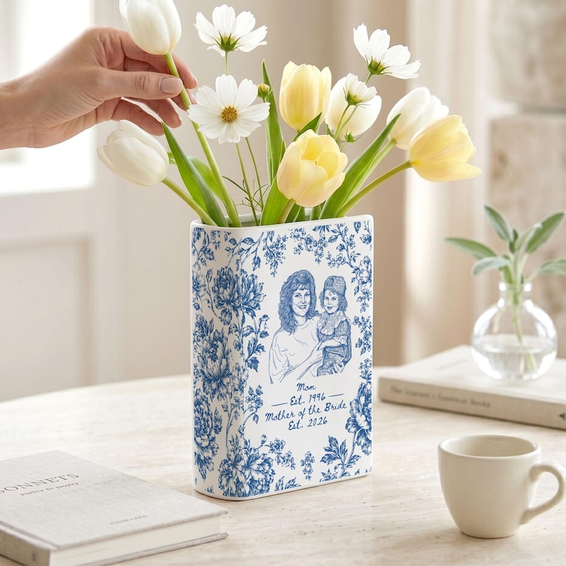 May include: A rectangular ceramic vase featuring a blue and white floral pattern. The front displays a line-art illustration of a person holding a child, with the text: Mom Est. 1996, Mother of the Bride Est. 2026. The vase holds white and yellow tulips.