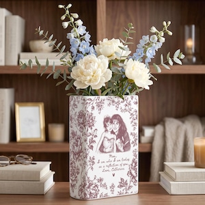 May include: A rectangular white ceramic vase featuring a maroon floral pattern and a printed illustration of two people. The text reads: Mom, I am who I am today as a reflection of you. Love, Catherine. The vase holds light blue and white flowers.