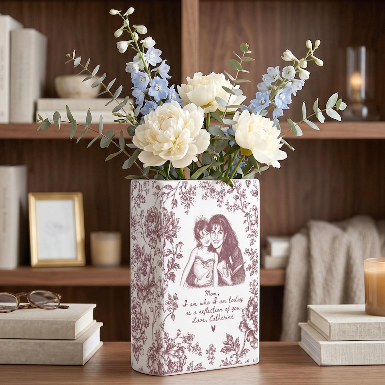 May include: A rectangular white ceramic vase featuring a maroon floral pattern and a printed illustration of two people. The text reads: Mom, I am who I am today as a reflection of you. Love, Catherine. The vase holds light blue and white flowers.