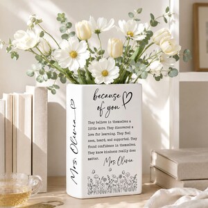 Puede incluir: Un florero rectangular de cer&aacute;mica blanca con texto en negro que dice because of you, Mrs. Olivia, y un p&aacute;rrafo sobre la bondad. El florero est&aacute; decorado con un patr&oacute;n floral negro en la base y lleno de flores blancas y hojas verdes.