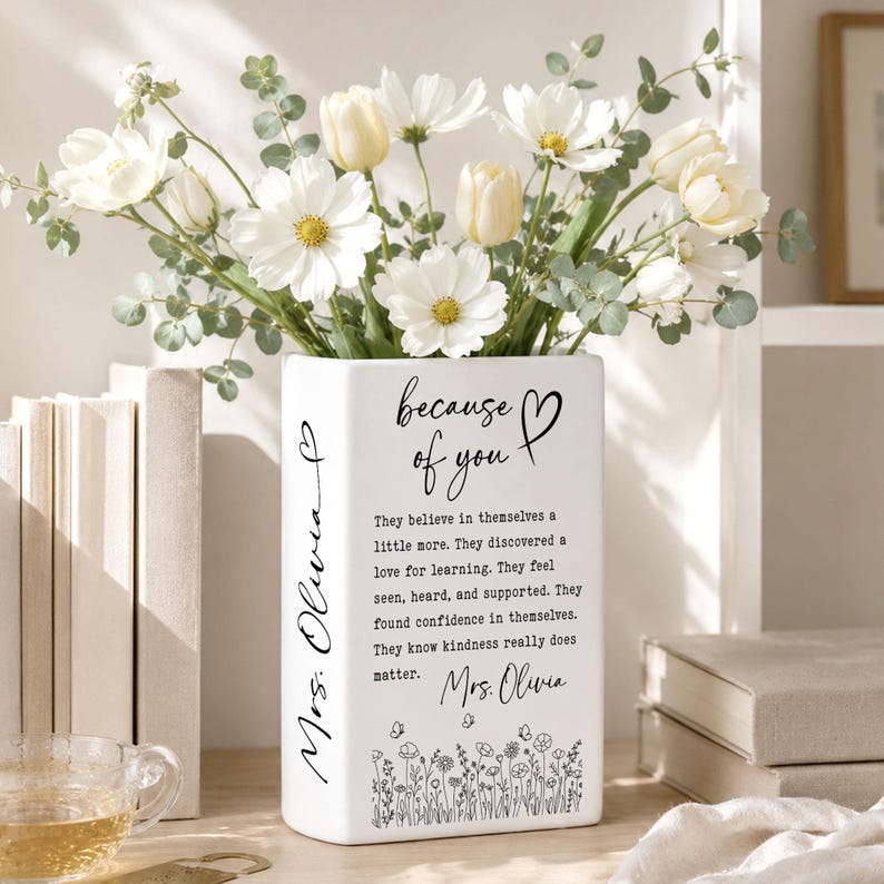 Puede incluir: Un florero rectangular de cer&aacute;mica blanca con texto en negro que dice because of you, Mrs. Olivia, y un p&aacute;rrafo sobre la bondad. El florero est&aacute; decorado con un patr&oacute;n floral negro en la base y lleno de flores blancas y hojas verdes.
