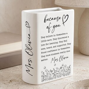 Puede incluir: Un florero rectangular de cer&aacute;mica blanca con texto en negro que dice because of you, seguido de un p&aacute;rrafo sobre la confianza y la amabilidad. El lateral est&aacute; personalizado con Mrs. Olivia y la base muestra un dise&ntilde;o floral de l&iacute;neas negras.