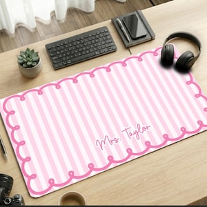 May include: A rectangular desk mat featuring a light pink and white vertical striped pattern with a dark pink scalloped border. The text "Mrs. Taylor" is printed in a cursive script font in the lower right section of the mat.