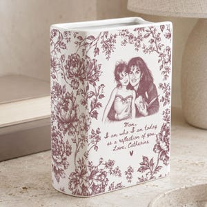 May include: A rectangular white ceramic vase featuring a dark pink floral pattern. The front displays a sketch of two people and the text: Mom, I am who I am today as a reflection of you. Love, Catherine, with a small heart symbol below the text.