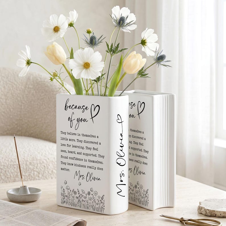 Puede incluir: Dos jarrones de cer&aacute;mica blanca con forma de libro que incluyen el texto negro "because of you" y "Mrs. Olivia" con un borde floral. Los jarrones contienen flores de cosmos blancas, tulipanes amarillos y tallos de cardo azul.