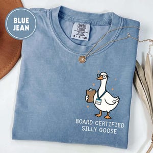 May include: Blue jean Comfort Colors t-shirt with a graphic of a goose wearing glasses and holding a clipboard. The text reads "BOARD CERTIFIED SILLY GOOSE". A gold necklace is draped on the shirt.