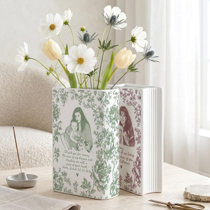 May include: Two rectangular white ceramic vases shaped like books, decorated with green and maroon floral patterns and custom portraits. The front vase features a green illustration of a person holding a baby with text below, while the second vase displays a maroon portrait and script text.
